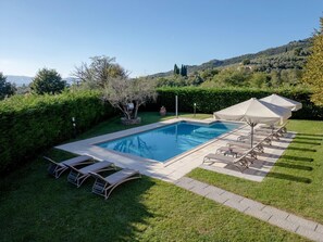 A rectangular swimming pool with a tiled deck, surrounded by a well-maintained lawn and a hedge. There are lounge chairs and a covered umbrella area.
