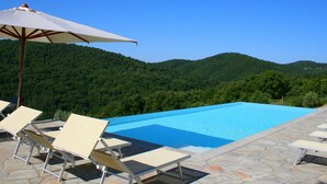 A swimming pool with lounge chairs and an umbrella, surrounded by stone tiles, with a forested mountain in the background.