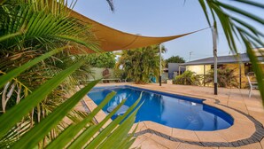 A swimming pool with a curved edge, surrounded by palm trees and a shaded area. - 79 Nimitz Street - Pool and Pet Friendly (Exmouth)