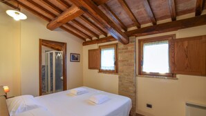 Une chambre avec un plafond en bois, deux fenêtres dotées de volets en bois, un lit avec du linge de maison blanc et une salle de bain avec une porte en verre.