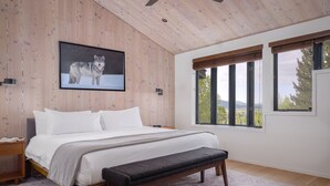 A bedroom with a bed, a bench, a picture of a wolf, and large windows with a view.