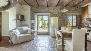 A cozy room with a stone wall, a sofa, a dining table with chairs, a television on the wall, and a refrigerator.
