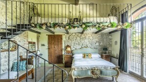 A bedroom with a stone wall, a bed with ornate headboard, a staircase with a wrought iron railing, and a view of the outdoors through a window.