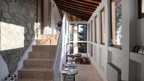 Un pasillo con paredes de piedra, vigas de madera y suelo de baldosas. Hay escalones que conducen a un rellano con una mesa y sillas.