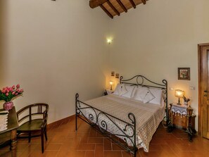 Une chambre à coucher avec un lit en métal, une chaise en bois, une petite table sur laquelle est posé un vase de fleurs et une porte en bois.