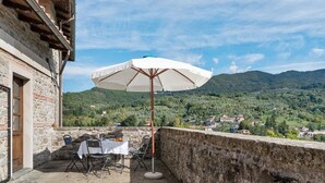 A stone building with a terrace featuring a table and chairs under a white umbrella, overlooking a mountainous landscape.