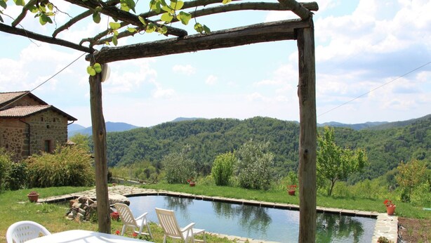 A rustic outdoor seating area with a table and chairs, a wooden pergola, and a view of a mountain landscape. - Holiday Home in Tuscany with Natural Pond (Mulazzo)