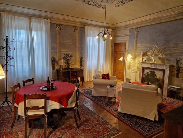 Ein Essbereich mit rotem Tischlein, Holzsesseln, einer Chandelier und einem Kamin mit einem Gemälde.