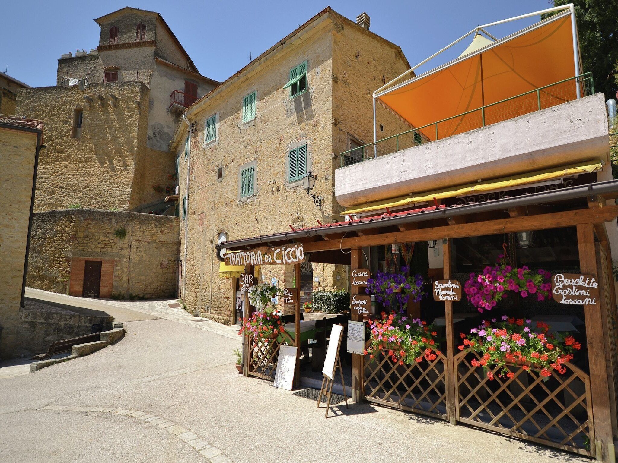 Un edificio de piedra tradicional con un bar llamado "Trattoria da Caccia" y una valla de madera adornada con cestas de flores colgantes.