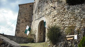 Un edificio in pietra con una torre e un portale ad arco, circondato da un'area verde e un muro in pietra.