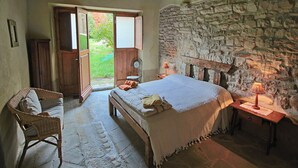 A bedroom with a stone wall, a bed with a white bedspread, a wicker chair, and a small table with a lamp.