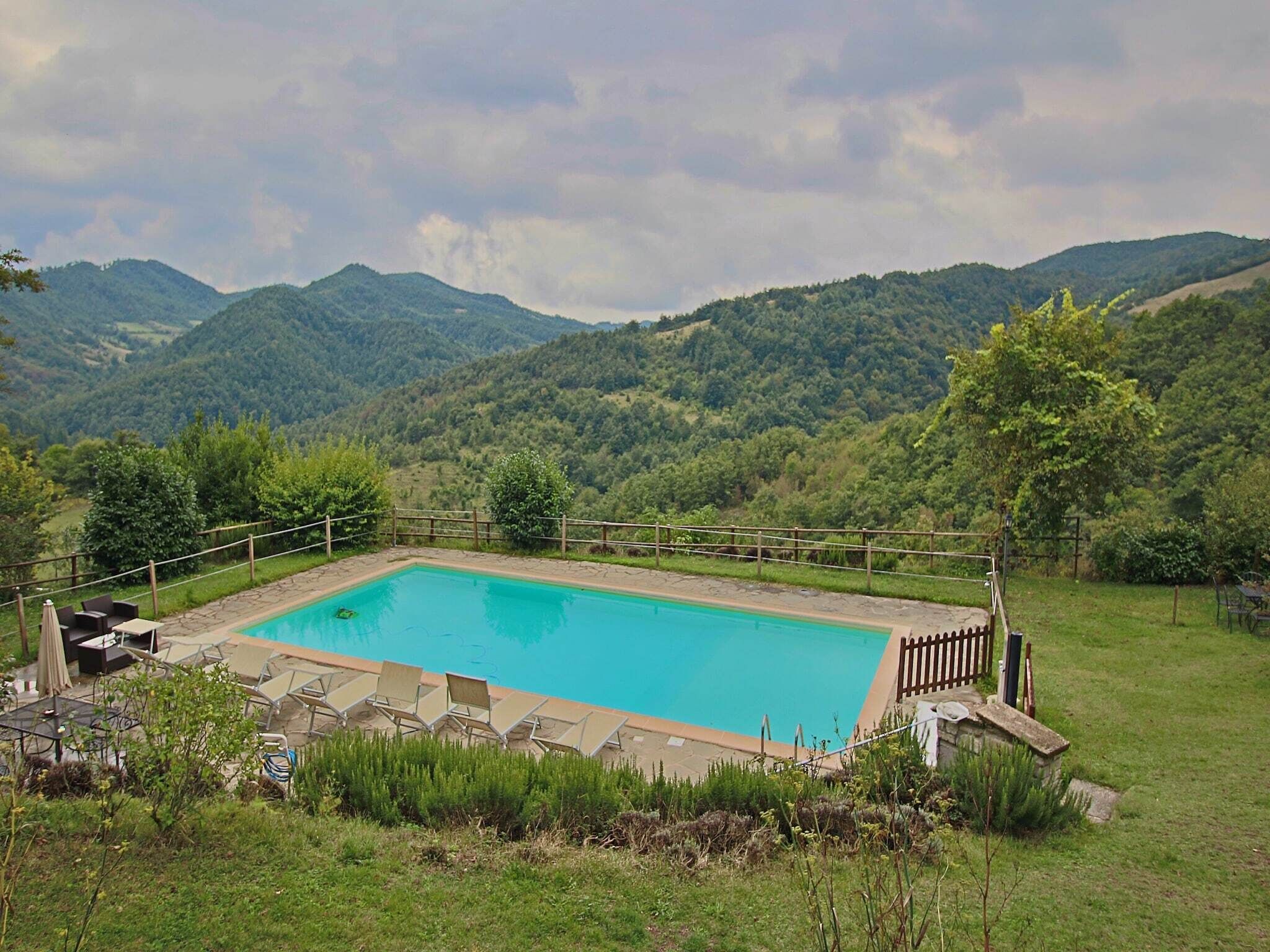 Ein rechteckiger Pool mit Holzterrasse und Liegestühlen, umgeben von einem Holzzaun, vor einer Kulisse aus üppigen grünen Hügeln und einem bewölkten Himmel.