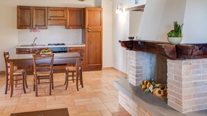 Une cuisine avec des meubles en bois, une table à manger, des chaises, une cheminée avec un manteau en briques et un luminaire suspendu.