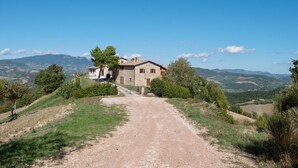 Une maison de campagne en pierre, accessible par un chemin de terre, entourée de verdure et avec des collines en arrière-plan.