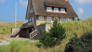 A two-story house with a steeply pitched roof, a staircase leading to the entrance, and a small garden area with a pine tree. - Villa in Midsland near the Sea (Midsland Terschelling)