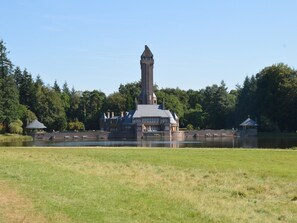 Un edificio grande e histórico con una torre destacada, ubicado junto a un lago con un primer plano de césped y árboles densos al fondo.
