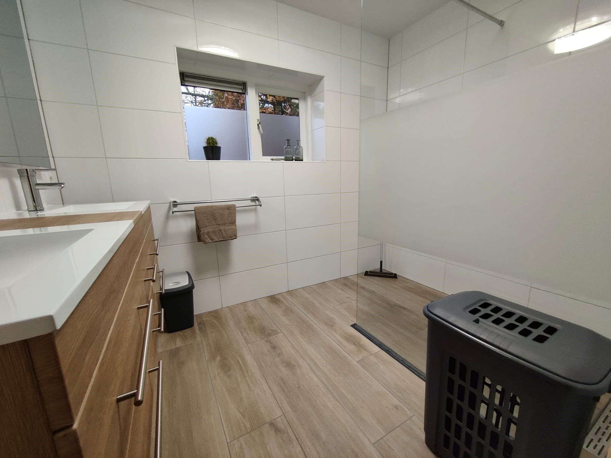 A modern bathroom with a wooden vanity, a towel rack, a trash bin, a mop, and a laundry basket.