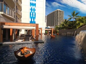 A hotel pool area with a waterfall, lounge seating, and a modern building in the background. - Waikiki | Couples Hawaii Beach Stay | Pool + Beach (Honolulu)