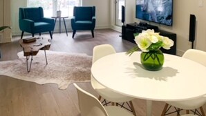 A modern living room with a round dining table, two blue armchairs, a white coffee table, and a large flat-screen TV mounted on the wall.
