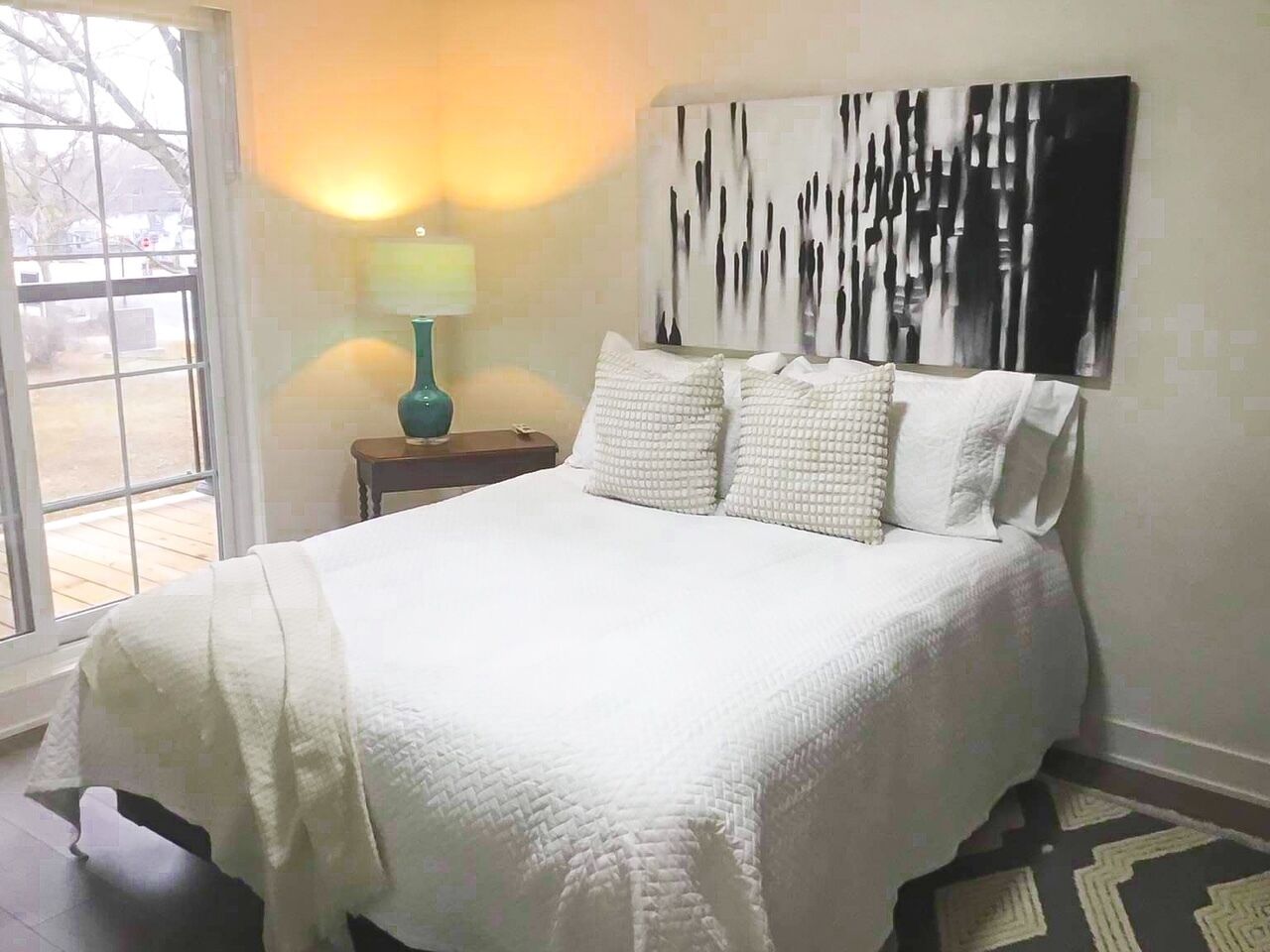 A bedroom with a large bed, a nightstand with a lamp, a window, and a wall with a black and white abstract painting.