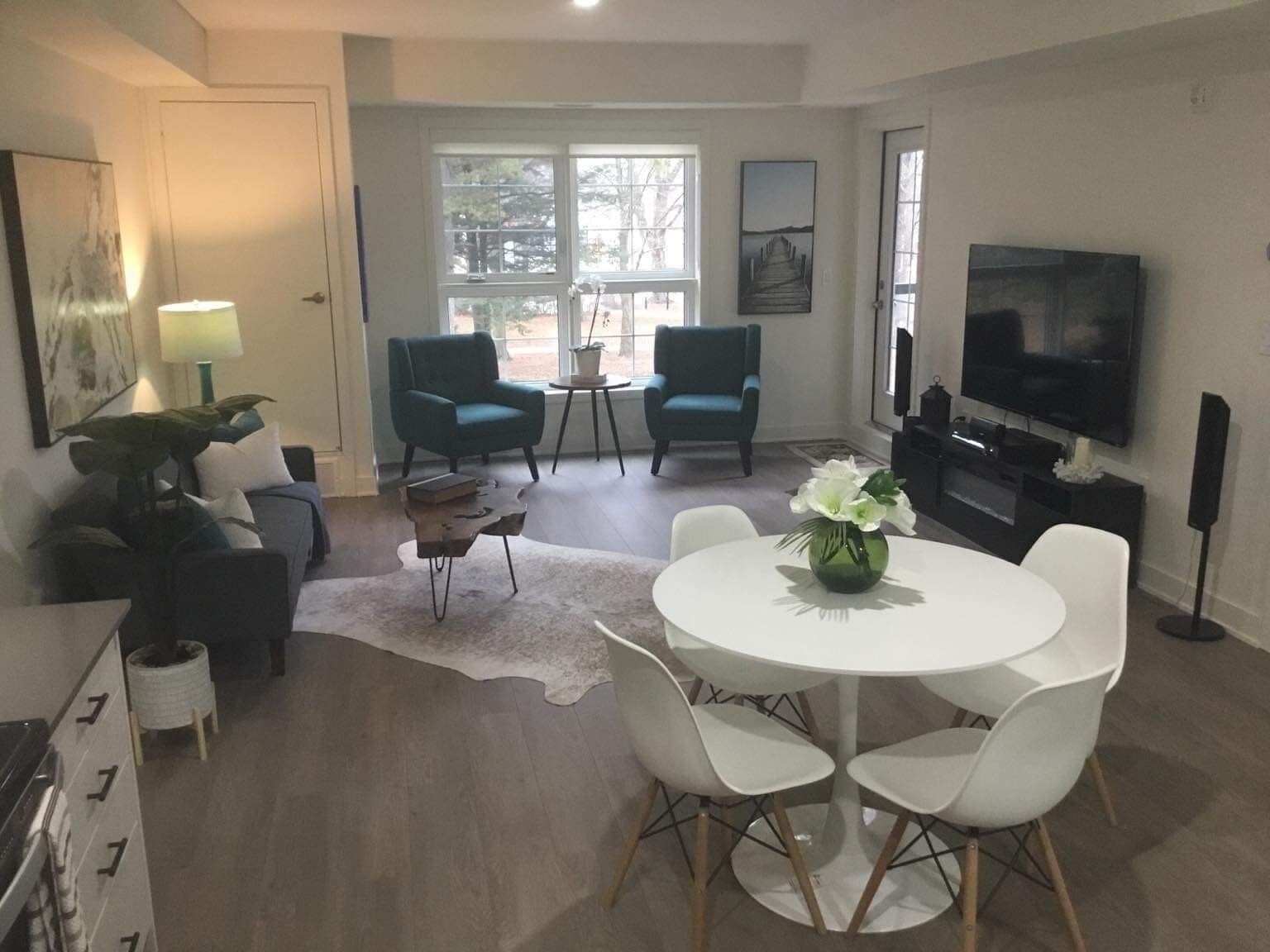 A modern living room with a round dining table, two chairs, a sofa, and two armchairs. There is a television and a speaker on the wall, and a window with a view of trees.