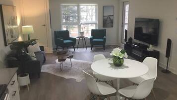 A modern living room with a round dining table, two chairs, a sofa, and two armchairs. There is a television and a speaker on the wall, and a window with a view of trees.