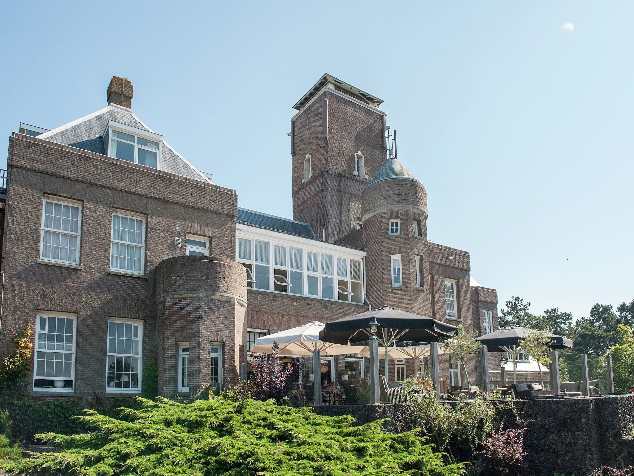 A large brick building with multiple windows and a tower. There are outdoor seating areas with umbrellas.
