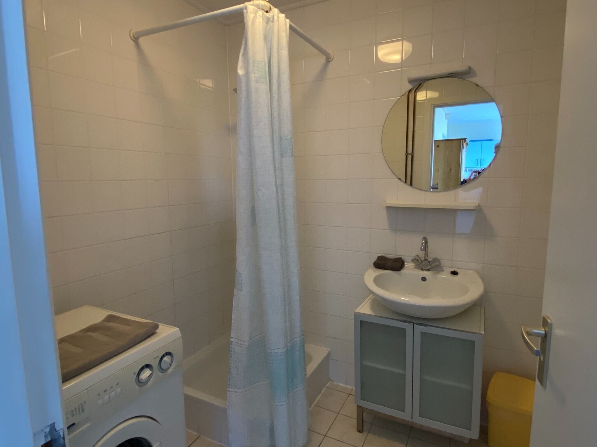 A bathroom with a shower, a washing machine, a sink, and a mirror.
