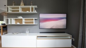 A modern living room with a flat-screen TV mounted on the wall, a white entertainment unit below, and a minimalist shelving unit with decorative items. - Holiday home near Grevelingen Lake (Zonnemaire)