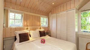 A bedroom with a wooden ceiling, a bed with white and brown bedding, two bedside lamps, a window with a view of greenery, and a white radiator.