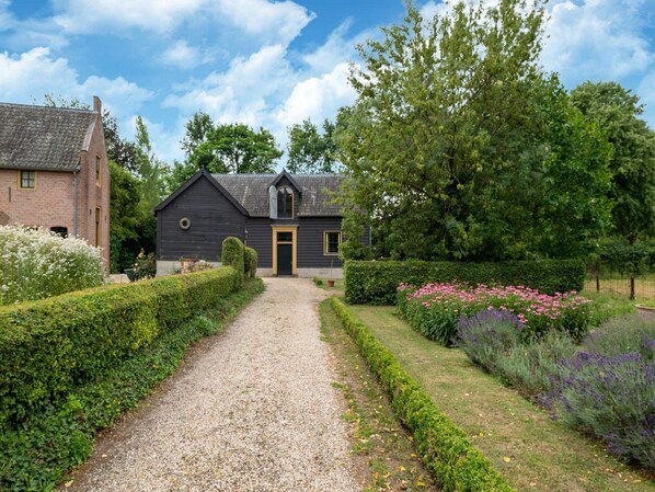 A gravel path leading to a dark wooden house with a yellow door, surrounded by well-maintained hedges and flowering plants. - Mansion in Deil by River Linge (Deil)