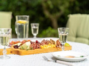 Una tabla de embutidos y quesos variados, acompañada de vasos de agua y una copa de vino blanco, sobre una mesa al aire libre.