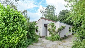 A small brick building with a sloped roof, surrounded by lush greenery and a stone pathway leading to it. - Lake Escape w/ Garden (Kamperland)