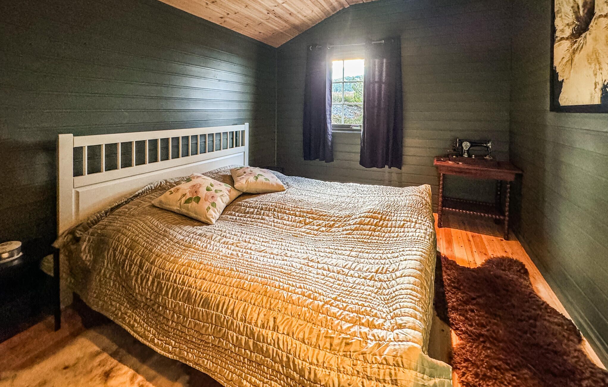 Un dormitorio acogedor con techo de madera, una cama grande con cabecera blanca, dos almohadas, una mesita de noche de madera, una máquina de coser antigua y una ventana con cortinas.