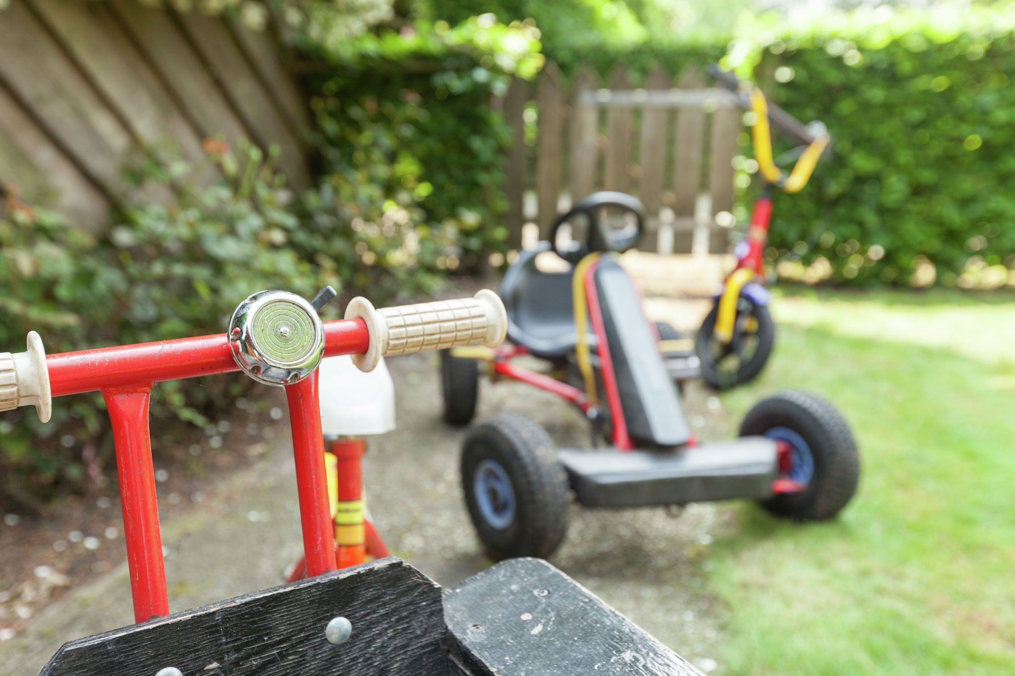 Un tricycle rouge, muni d’un guidon et d’un siège, est garé dans un jardin. On aperçoit une clôture en bois et de la verdure en arrière-plan.