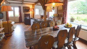 A dining area with a wooden table, chairs, and a chandelier. There is a television in the corner and a window with curtains. - Hideaway: Sauna, Tennis & Trampoline (Oploo)