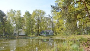 A serene lakeside setting with two houses, one with a green roof and another with a white facade, surrounded by lush greenery and trees.