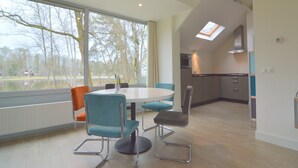 A modern interior with a dining area featuring a round table and teal chairs, a kitchen with a skylight, and a view of trees through large windows.