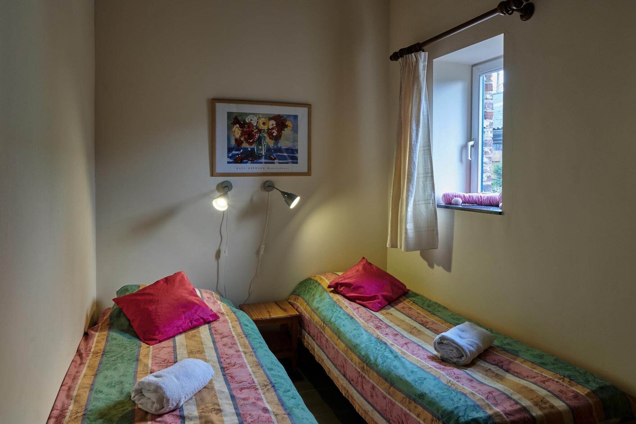 A small room with two single beds, each with a colorful striped bedspread and a red pillow. There is a small table between the beds, a wall-mounted lamp, and a window with a curtain.