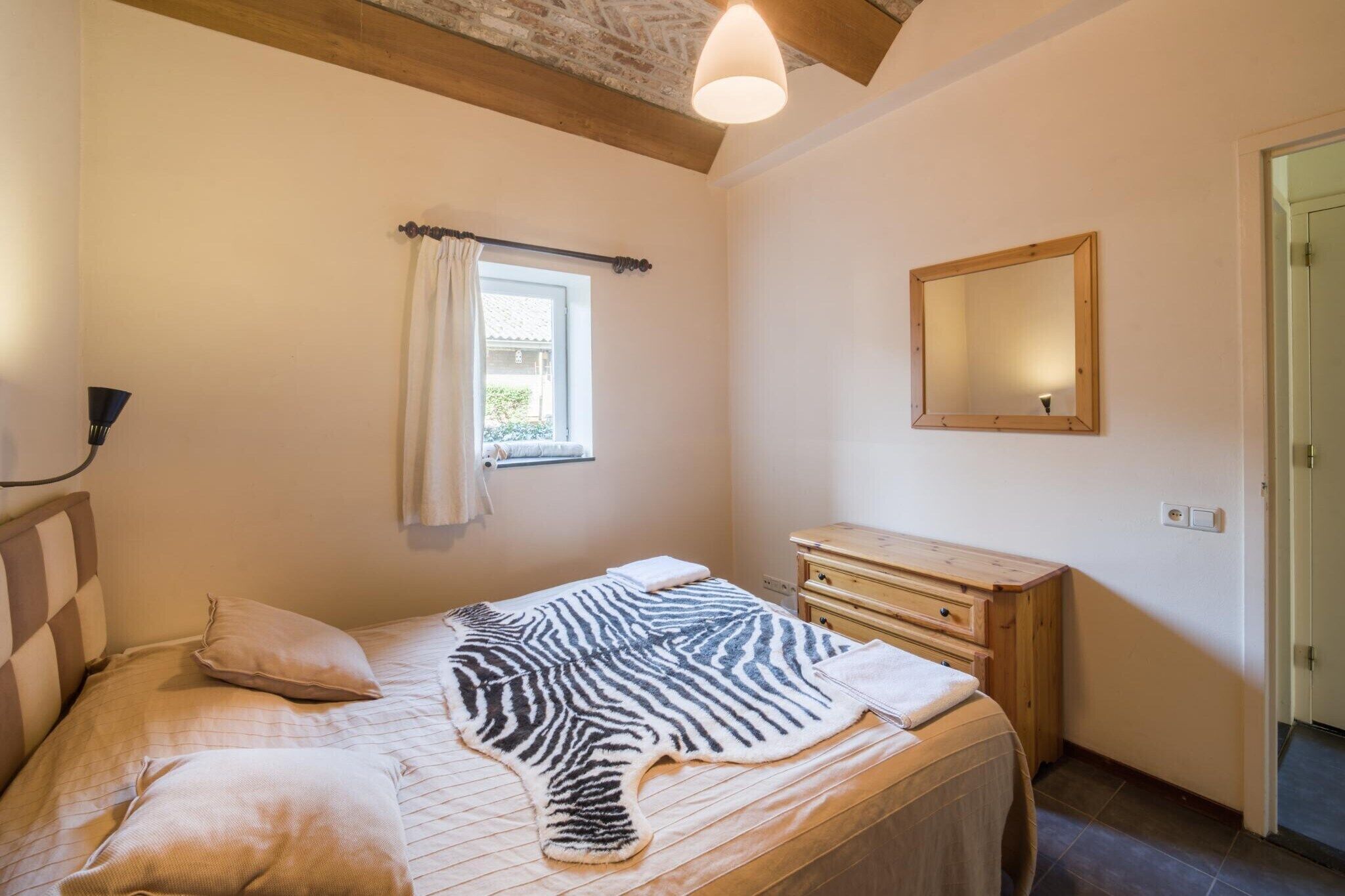 A bedroom with a zebra-print bedspread, wooden headboard, a bedside table, a mirror, and a window with curtains.