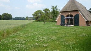 Un bâtiment traditionnel à toit de chaume, avec une façade en briques, un petit patio doté de sièges extérieurs et, au premier plan, un champ de fleurs blanches.