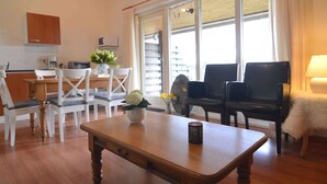 Un salon chaleureux avec une table basse en bois, deux chaises noires, une table à manger blanche avec des chaises, et un coin cuisine doté de meubles en bois et d’une fenêtre avec des stores.