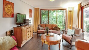 Een woonkamer met een bank, fauteuils, een salontafel, een tv op een houten kast en een groot raam met gordijnen.