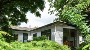 Een klein, laag huis met een witte gevel en groene accenten, omringd door weelderige begroeiing.
