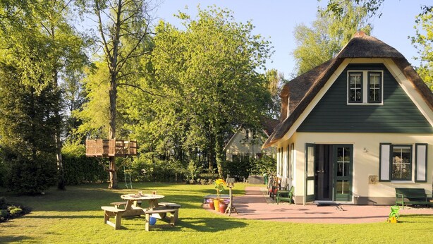 Una casa tradizionale con tetto di paglia, immersa in un giardino curato con tavoli da picnic e un'area giochi in legno.
