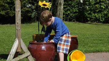 Un enfant joue avec un grand pot de fleurs et un seau jaune sur un chemin pavé.