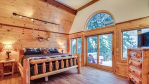 Un dormitorio de cabaña de madera con una cama grande, paredes de madera, una cabeza de ciervo montada y vistas de un paisaje nevado a través de amplias ventanas.