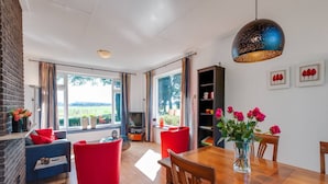 Una sala moderna con una mesa de comedor de madera, sillas rojas y azules, un televisor y un ventanal con vistas a zonas verdes.