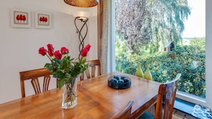 Un comedor con una mesa de madera, sillas, un jarrón con rosas rojas, una lámpara colgante y vistas de un jardín a través de un ventanal grande.