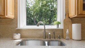 A kitchen with a stainless steel sink, wooden cabinets, a window with a view of trees, and a paper towel dispenser. - Tulsa Turnaround 4 bed (Tulsa)